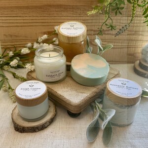 May include: A collection of self-care products, including a candle, soap, and jars of scrubs and creams. The candle is in a glass jar with the text "Sabbath Glow". The soap is a swirl of green and peach. The jars have wooden lids.