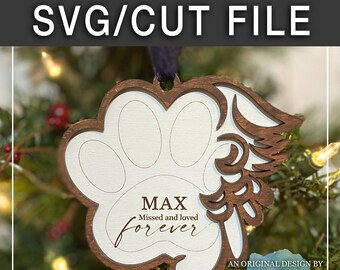 Op de afbeelding: Een houten poot afdruk ornament met een bruine vlek en witte verf. Het ornament heeft een uitgesneden ontwerp van een vleugel en de tekst "MAX Missed and loved forever". Het ornament hangt aan een kerstboom.