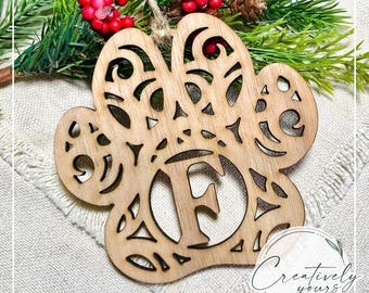 May include: A wooden paw print ornament with the letter "F" cut out in the center. The ornament has decorative cutouts and a twine loop for hanging. The background includes greenery and red berries. The ornament is from "Creatively Yours by DWS."