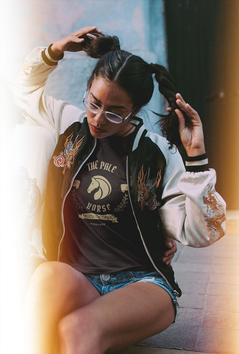 K&ouml;nnte beinhalten: Eine Frau tr&auml;gt ein schwarzes T-Shirt mit einer goldenen Grafik, auf der "The Pale Horse Much Deeping England" steht, und eine wei&szlig;-schwarze Bomberjacke mit einem gestickten Drachenmuster. Sie tr&auml;gt Jeans-Shorts und hat ihre Haare zu zwei Pferdeschw&auml;nzen gebunden.