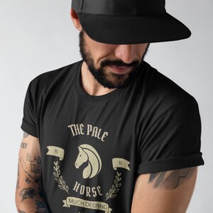 K&ouml;nnte beinhalten: Ein schwarzes T-Shirt mit einem Grafikdesign, das einen wei&szlig;en Pferdekopf mit dem Text "The Pale Horse" und "Much Deeping" in einem Vintage-Stil zeigt.