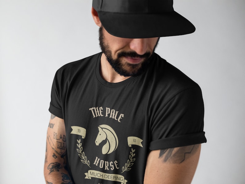 K&ouml;nnte beinhalten: Ein schwarzes T-Shirt mit einem Grafikdesign, das einen wei&szlig;en Pferdekopf mit dem Text "The Pale Horse" und "Much Deeping" in einem Vintage-Stil zeigt.