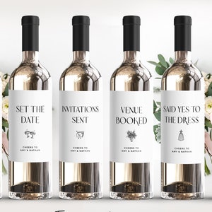 May include: Six white wine bottles with black labels. Each label has a different engagement-themed phrase and icon, such as "Honeymoon Booked", "Set the Date", "Invitations Sent", "Venue Booked", "Said Yes to the Dress", and "Cake Tasting". The labels also include the text "Cheers to Amy & Nathan".