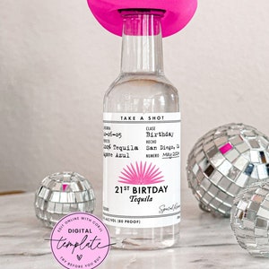 May include: A clear glass bottle of tequila with a pink cowboy hat on top. The label reads "21st Birthday Tequila" with a pink sunburst design. The bottle is surrounded by silver disco balls.
