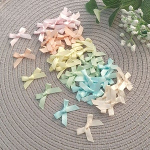 May include: A collection of pastel colored satin ribbon bows. The bows are arranged in a rainbow pattern, starting with pink and ending with white. The bows are on a gray textured surface.
