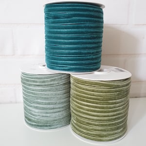 May include: Three spools of velvet ribbon in teal, sage green, and olive green. The ribbon is wound around white spools.