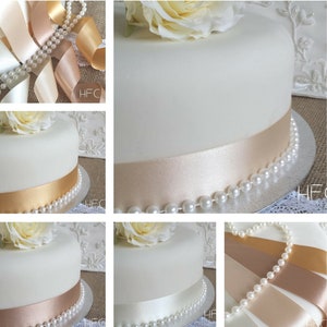 May include: A white cake decorated with a single white rose and a beige satin ribbon. The cake is on a silver cake stand with a pearl trim.