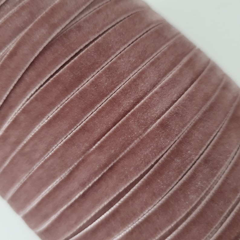 May include: A close-up of a roll of dusty rose velvet ribbon. The ribbon is folded in a zig-zag pattern, showing the soft texture of the fabric.