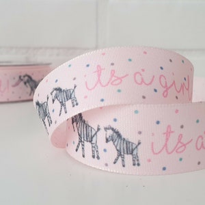 May include: Pink ribbon with a zebra print and the text "it's a girl" in pink. The ribbon is decorated with small, colorful confetti.
