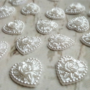 May include: A collection of white pearl heart-shaped embellishments with a raised rose design. Each heart has a beaded border.