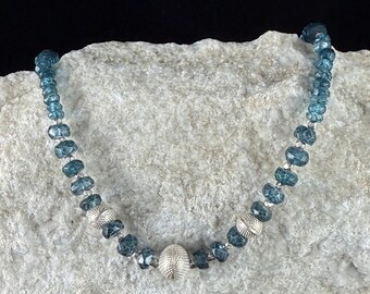 May include: A blue beaded necklace with silver accents. The necklace features faceted blue beads interspersed with silver-colored, textured beads and a larger, textured silver bead at the center. The necklace is displayed on a light-colored, textured surface.