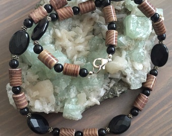 May include: A necklace made of black and brown wooden beads with a silver clasp. The necklace is displayed on a bed of green and white crystals.