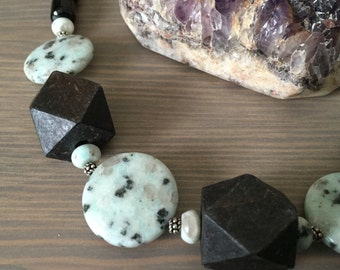 May include: A necklace with black, white, and green beads. The beads are made of various materials, including wood, stone, and metal. The necklace is on a wooden surface.