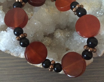 May include: A bracelet made with carnelian and black beads. The carnelian beads are round and orange-red in color. The black beads are round and shiny.