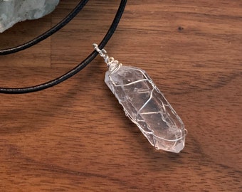 May include: A clear quartz crystal pendant wrapped in silver wire, hanging from a black leather cord.