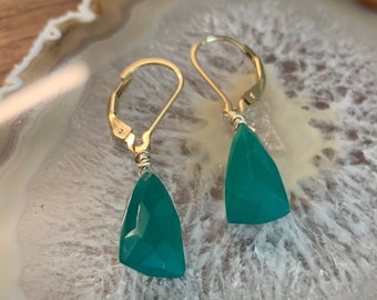 May include: A pair of gold earrings with green gemstone teardrop dangles. The earrings are on a white and brown agate slice.