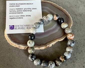 May include: A gray, black, and white jasper stone bracelet with a silver charm. The bracelet is on a gray and white agate coaster. The text on the card reads "POSITIVE RELATIONSHIPS BRACELET picasso jasper PROMOTES meditation, grounding, peace, harmony, positive relationships, emotional balance PROTECTS AGAINST stress, anxiety, negative emotions WWW.TRACYKREIFELSA WEARABLE HEAI"