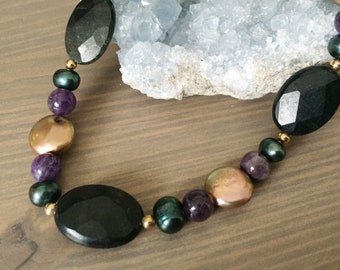 May include: A necklace made with black and green gemstone beads, gold beads, purple amethyst beads, and iridescent brown beads.