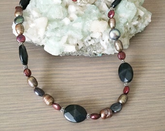 May include: A long necklace made of black oval beads, brown and gray pearl beads, and small red beads.