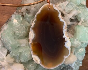 May include: A brown agate pendant with a white edge, hanging from a copper wire necklace. The pendant is resting on a bed of green and white crystals.