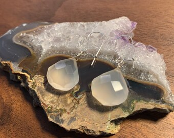 May include: A pair of silver earrings with white chalcedony gemstone drops. The earrings are on a natural agate geode.