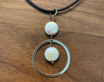 May include: A silver necklace with a black leather cord and a pendant featuring two white pearls and a silver circle.