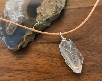 May include: A clear quartz crystal pendant wrapped in silver wire, suspended from a tan leather cord. The necklace is displayed on a dark wood surface, with a large, multi-toned agate slice in the background. The crystal is approximately 2 inches long.