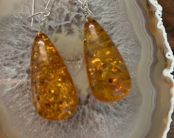 May include: A pair of amber teardrop earrings with silver hooks. The amber is a deep golden color with flecks of gold throughout.