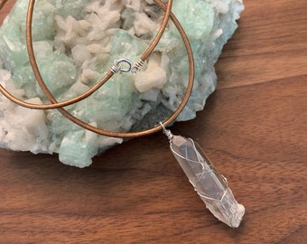 May include: A clear quartz crystal wrapped in silver wire hangs from a brown leather cord necklace. The necklace is displayed on a bed of green and white crystals.
