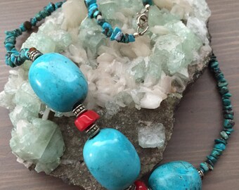 May include: A turquoise and red beaded necklace with large turquoise beads and silver accents. The necklace is displayed on a bed of green and white crystals.