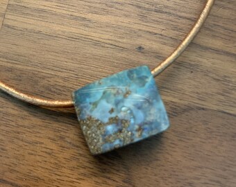 May include: A square-shaped pendant with a blue and white marbled design and gold flecks, hanging from a brown leather cord.