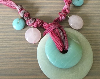 May include: A pink and green knotted fabric necklace with a large green jade pendant. The pendant is made of two overlapping circles. The necklace also features smaller pink and green beads.