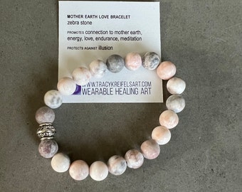 May include: A bracelet made of pink and gray zebra stone beads with a silver charm. The bracelet is on a white card with the text "MOTHER EARTH LOVE BRACELET zebra stone PROMOTES connection to mother earth, energy, love, endurance, meditation PROTECTS AGAINST illusion ...WW.TRACYKREIFELSART.COM WEARABLE HEALING ART".