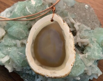May include: A natural stone pendant necklace. The pendant is a polished, oval-shaped agate with a cream and tan color scheme. The stone is suspended from a light brown cord, with copper wire detailing at the top. The necklace rests on a light green mineral formation.