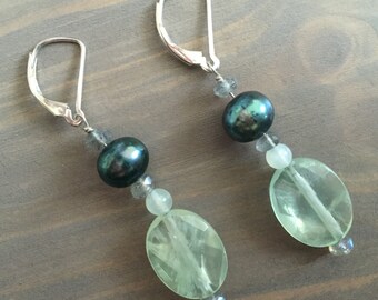 May include: A pair of dangle earrings featuring a dark teal pearl, light green oval stone, and small clear beads. The earrings have silver-colored lever-back closures and are displayed on a wooden surface.