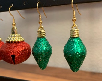 May include: A pair of green glitter Christmas light bulb earrings and a pair of red glitter bell earrings. The earrings are hanging from a black display stand.