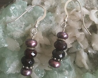May include: A pair of silver dangle earrings with a stack of dark purple and burgundy beads. The beads are faceted and have a metallic sheen.