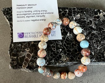 May include: A tranquility bracelet made with impression jasper beads. The bracelet is brown, blue, and white with a silver charm. The bracelet is on a black and white surface.  "TRANQUILITY BRACELET impression jasper PROMOTES bonding, unifying energy. encouragement, physical and emotional recovery, alignment, tranquility PROTECTS AGAINST negative energy WWW.TRACYKREIF WEARABLE HE COM ART"