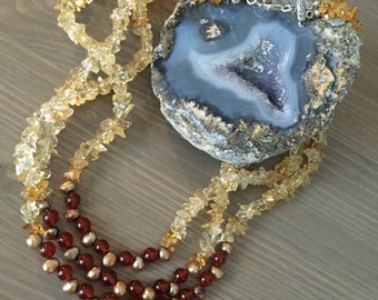 May include: A multi-strand necklace with brown and gold beads. The necklace is made of carnelian and citrine stones. The necklace is displayed on a slice of agate.