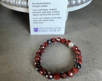 May include: A self-healing bracelet made with mahogany obsidian beads and a silver charm. The bracelet is on a gray surface with a card that reads "SELF-HEALING BRACELET mahogany obsidian PROMOTES self-healing, meditation, self-control, restful sleep, emotional balance, spiritual growth, grounding PROTECTS AGAINST allergies, psychic attack, stress, grief, negative energy. energy blocks WWW.TRACYKREIFELSART.COM WEARABLE HEALING ART"