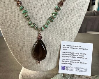 May include: A brown smoky quartz crystal pendant necklace with turquoise and clear glass beads. The necklace is displayed on a fabric-covered mannequin. The card below the necklace reads "Joy & Friendship Necklace turquoise, smoky quartz, sterling silver, crystal Promotes spirituality, clarity, friendship, manifestation of dreams, balance, grounding, meditation, awareness, joy Protects against mental chaos, depression, tension, anger, resentment, negative energy WWW.TRACYKREIFELSART.COM WEARABLE HEALING ART"