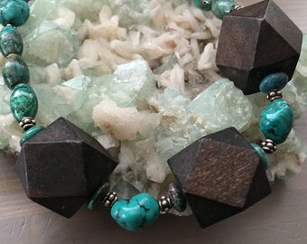May include: A turquoise and wood bead necklace with large, dark brown, geometric wooden beads. The necklace is displayed on a bed of light green and white crystals.