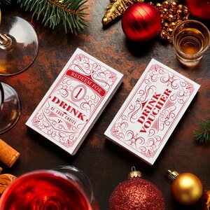 May include: Two white drink vouchers with red and gold ornate designs. The voucher on the left says "Redeem for 1 Drink of your choice". The voucher on the right says "Drink Voucher". The vouchers are on a brown surface with red and gold Christmas ornaments.