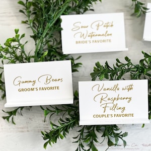 May include: Three white rectangular place cards with gold foil lettering. The cards read Sour Patch Watermelon BRIDE'S FAVORITE, Gummy Bears GROOM'S FAVORITE, and Vanilla with Raspberry Filling COUPLE'S FAVORITE. Each card has a folded base.