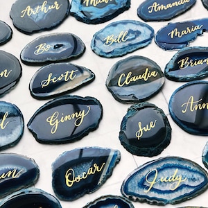 May include: A collection of blue agate slices with gold calligraphy names, such as "Arthur", "Michael", "Alexandra", "Amanda", "Bill", "Maria", "Scott", "Claudia", "Brian", "Ginny", "Ann", "Sue", "Oscar", "Judy", "Mitch", and "Jeanne".