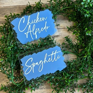 May include: Two acrylic signs with blue painted backgrounds and white text. The top sign says "Chicken Alfredo" and the bottom sign says "Spaghetti".