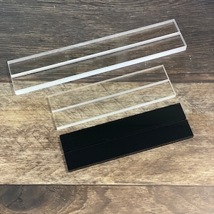 May include: Three acrylic display stands, two clear and one black. The stands are made of clear acrylic and have two slots for displaying items. The black stand is made of black acrylic and has two slots for displaying items.