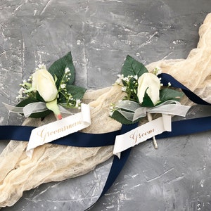 May include: Two white flower boutonnieres with green leaves and white ribbon. One boutonniere has a white tag with the word "Groom" written in gold script. The other boutonniere has a white tag with the word "Groomsman" written in gold script.