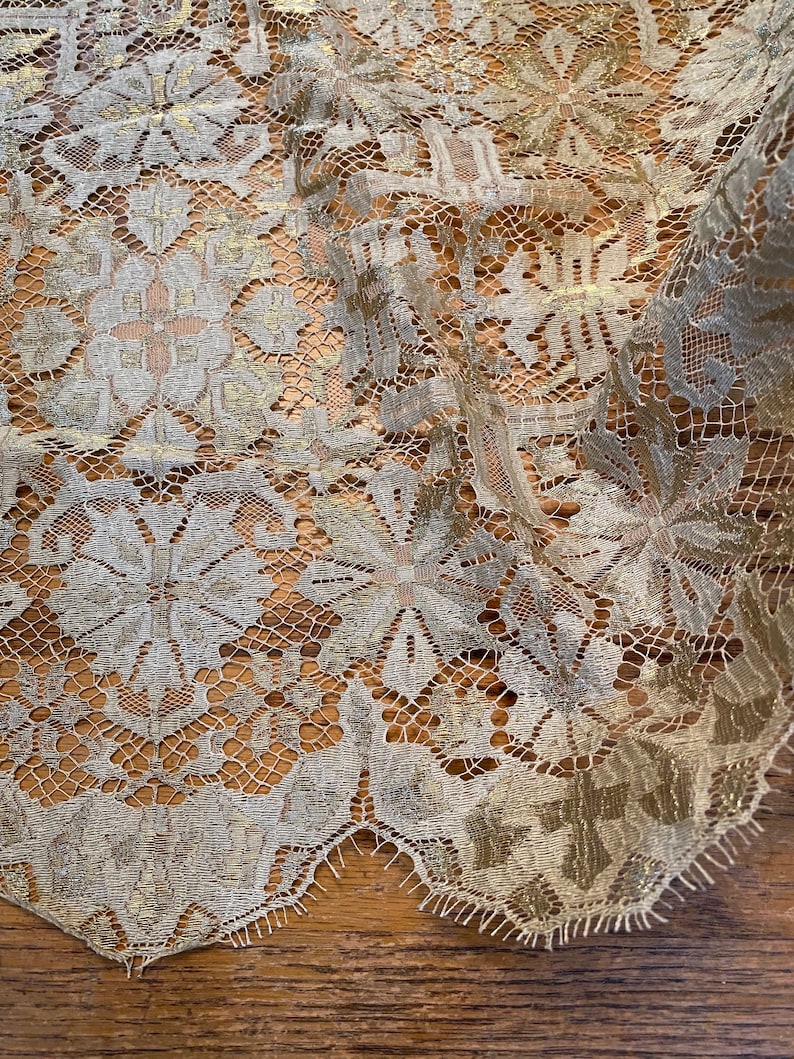 May include: A close-up of a gold and white lace fabric with a floral pattern. The lace is delicate and intricate, with a slightly textured surface.
