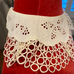Puede incluir: Collar de encaje blanco con borde festoneado y delicado bordado floral. El collar est&aacute; sobre un fondo de terciopelo rojo.